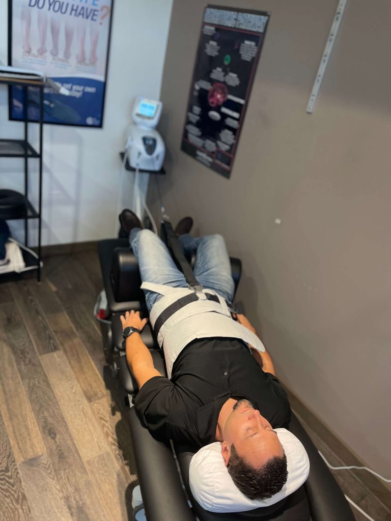 Picture showing a male patient laying on a medical bed. He has a spinal decompression machine attached to his torso. He wears a black shirt and blue jeans.