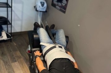 Picture showing a male patient laying on a medical bed. He has a spinal decompression machine attached to his torso. He wears a black shirt and blue jeans.